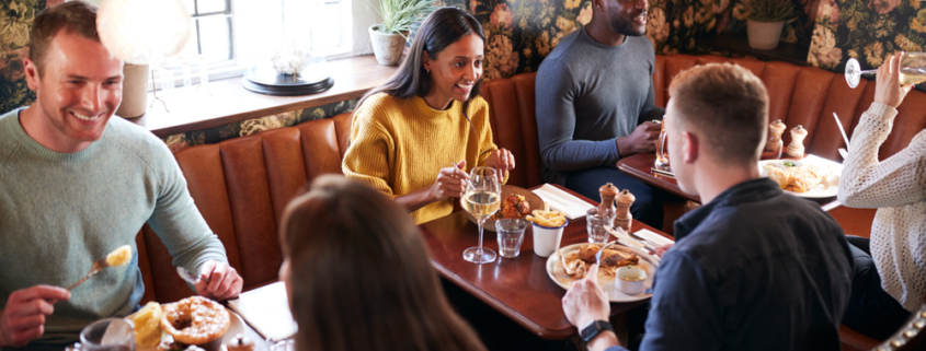 Group of friends enjoying a meal together inside a warmly decorated restaurant with booth seating.